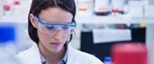 A woman in a lab wearing safety glasses and a lab coat looks down.