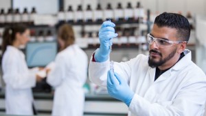 A man in a lab wearing safety glasses, gloves, and a lab coat examines a vial of water.