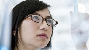 A woman with brown hair and glasses holds a test tube in the air to examine it.